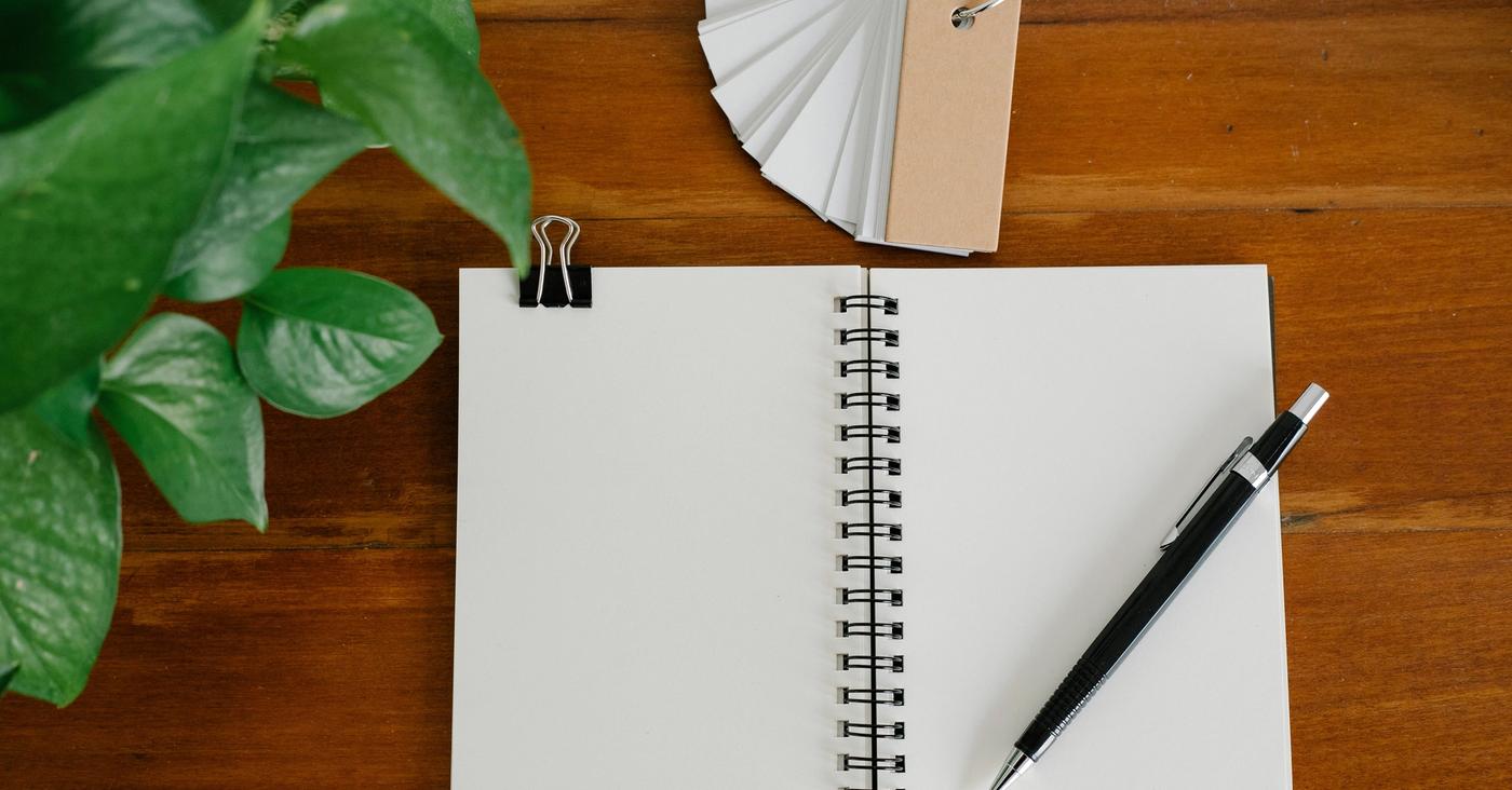 Open blank notebook and pen beside paper tags and a green plant on a wooden desk.