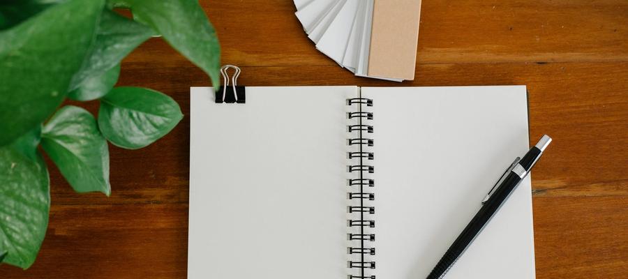 Open blank notebook and pen beside paper tags and a green plant on a wooden desk.