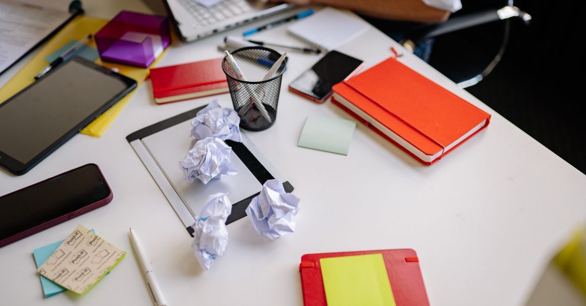 Crumpled papers, notebooks and stationery cover a desk.