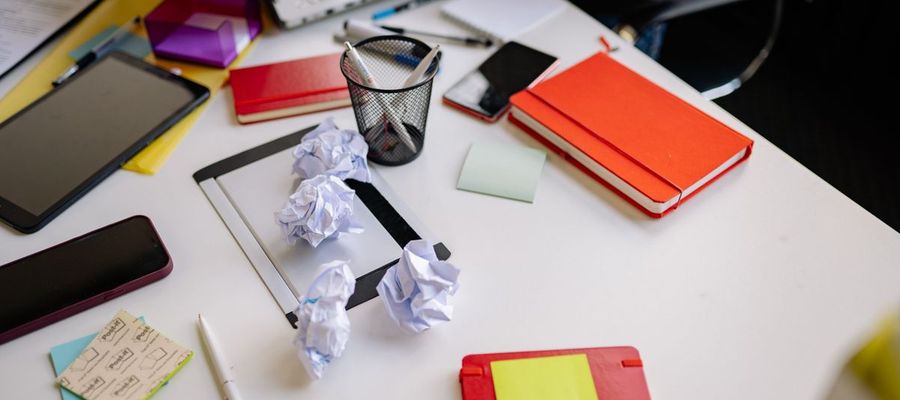Crumpled papers, notebooks and stationery cover a desk.