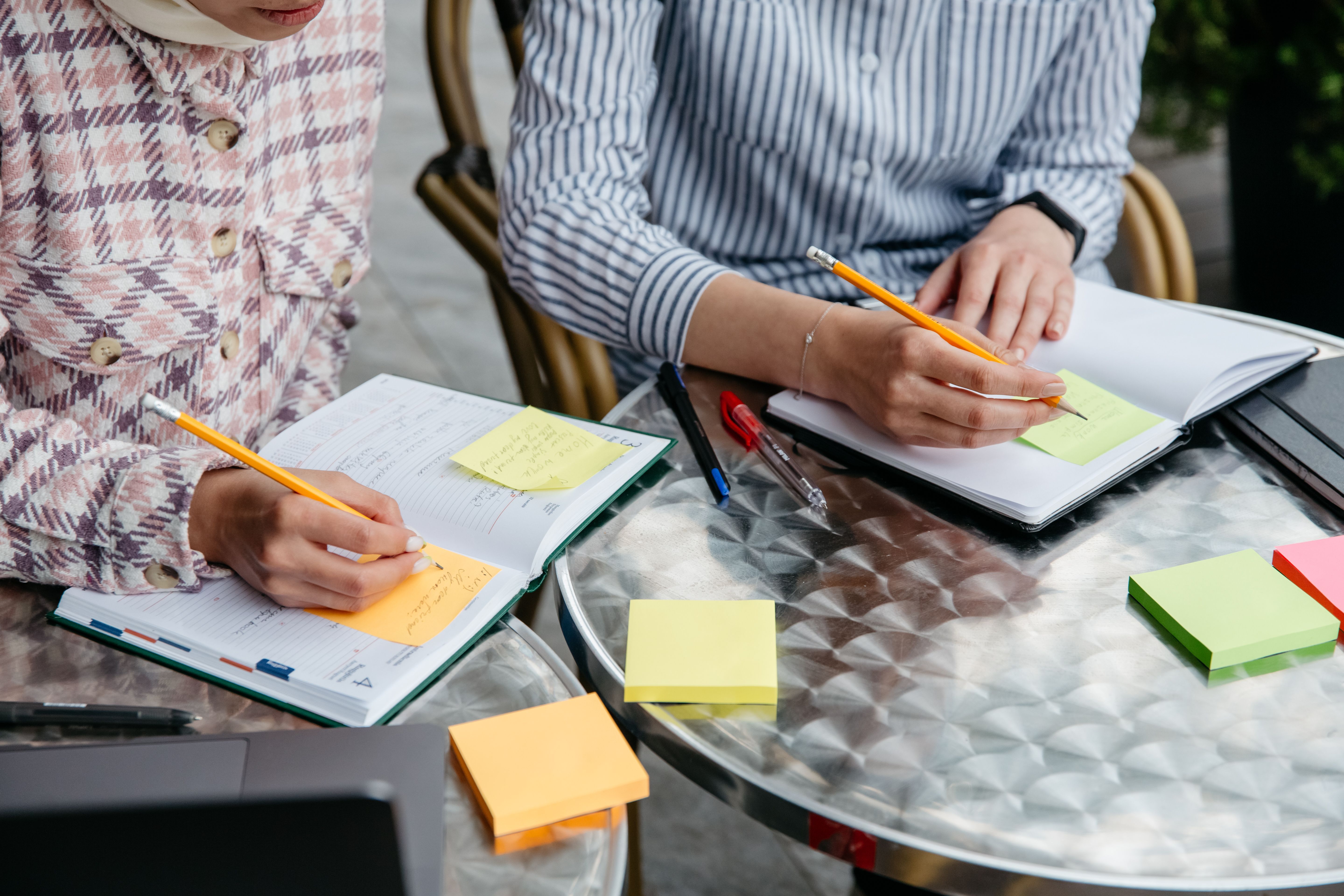 Two people writing in notebooks with colorful sticky notes on a shared table.