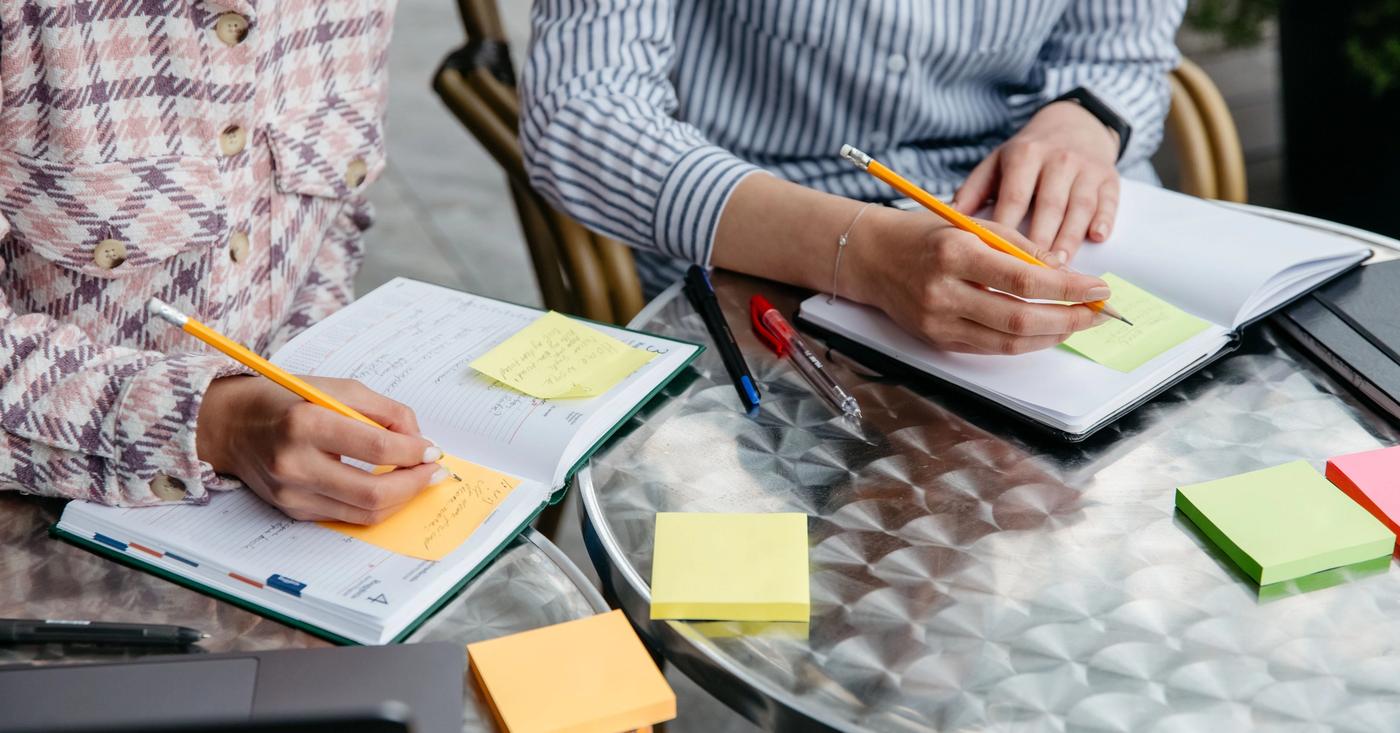 Two people writing in notebooks with colorful sticky notes on a shared table.