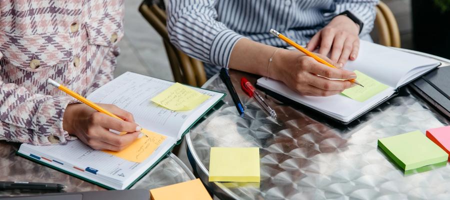 Two people writing in notebooks with colorful sticky notes on a shared table.