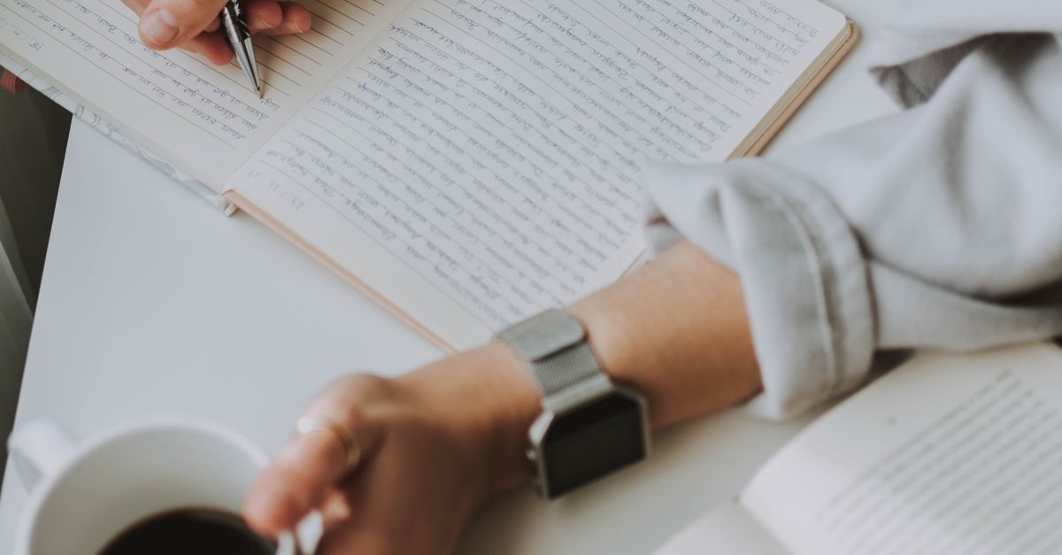 A person writes in a notebook while holding a coffee mug.
