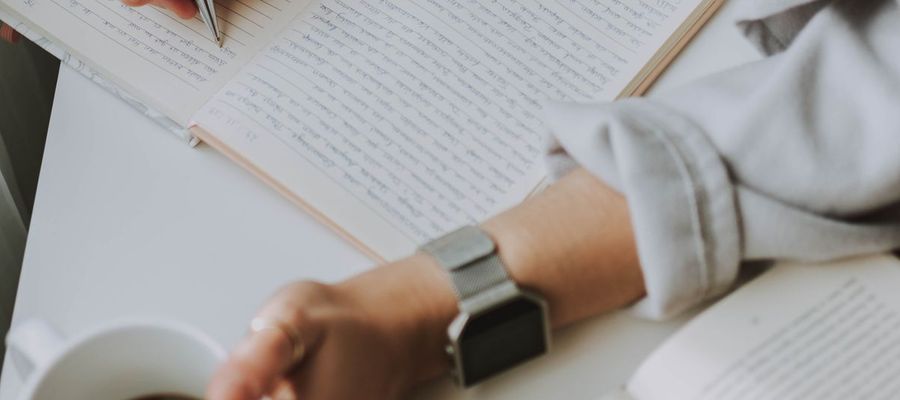 A person writes in a notebook while holding a coffee mug.
