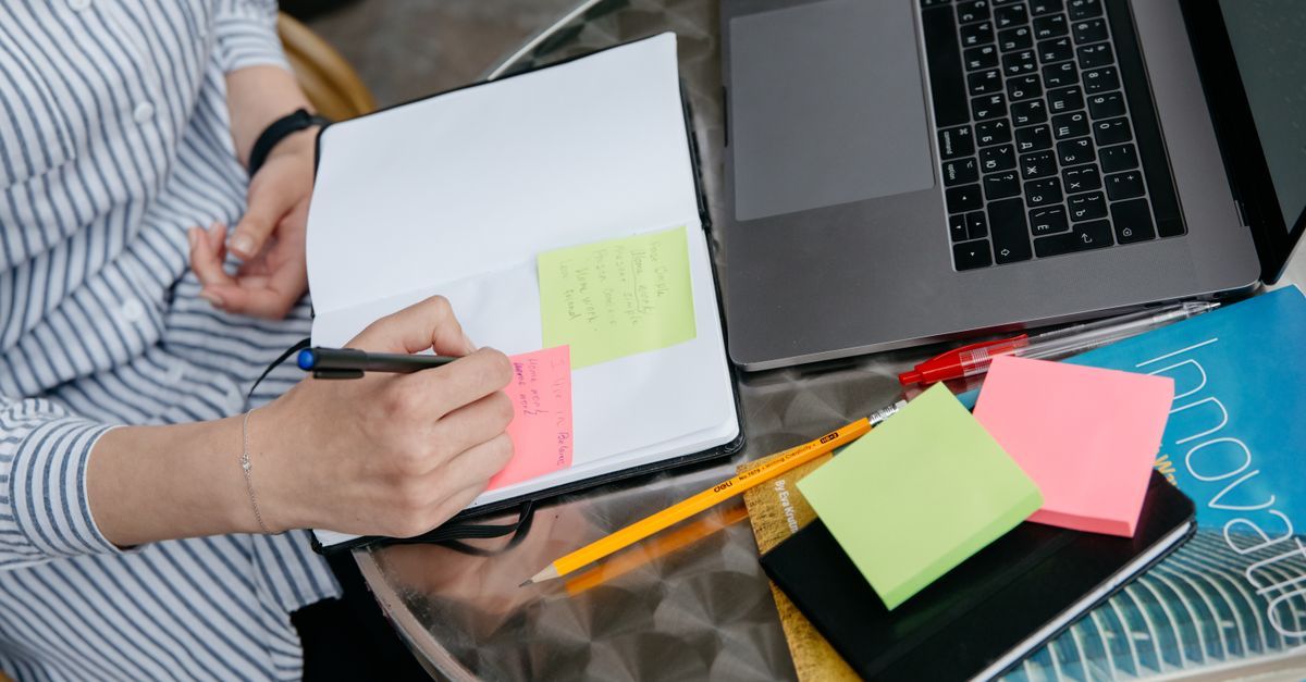 A person writes in a notebook beside a laptop and sticky notes.