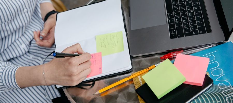 A person writes in a notebook beside a laptop and sticky notes.