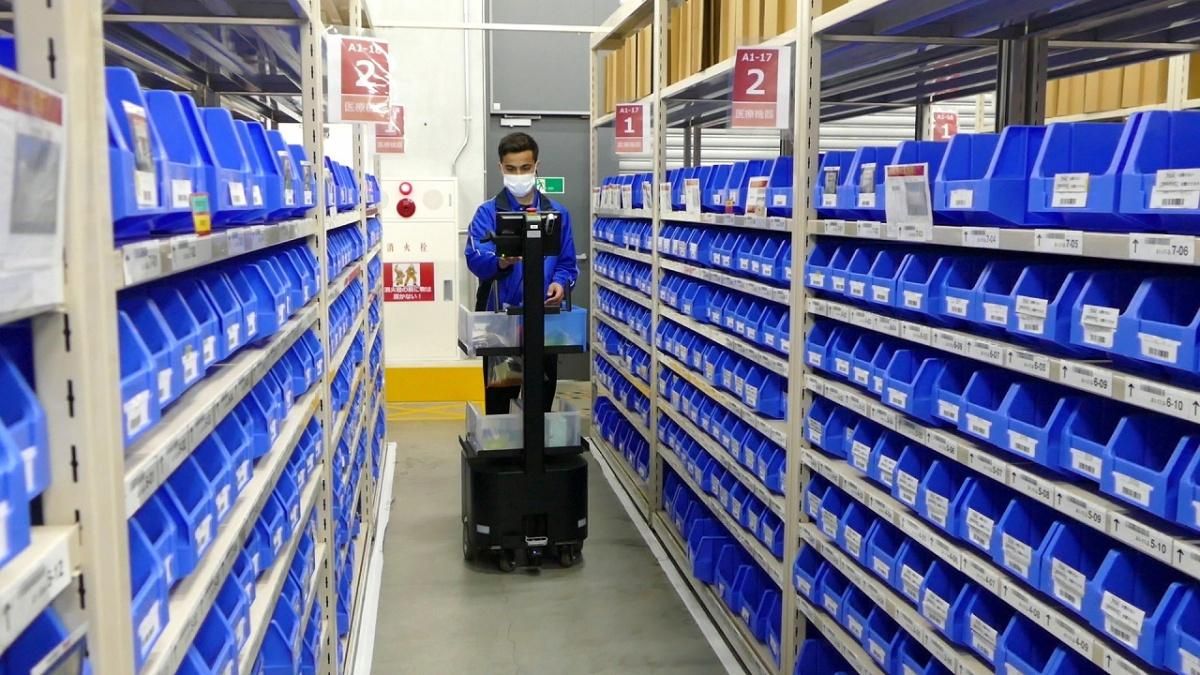 Warehouse worker picking from blue-bin shelves beside a mobile picking robot