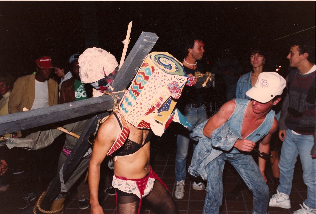 Border Arts Workshop performer wearing ladies lingerie, covered in a mask, and carrying a crucifix.
