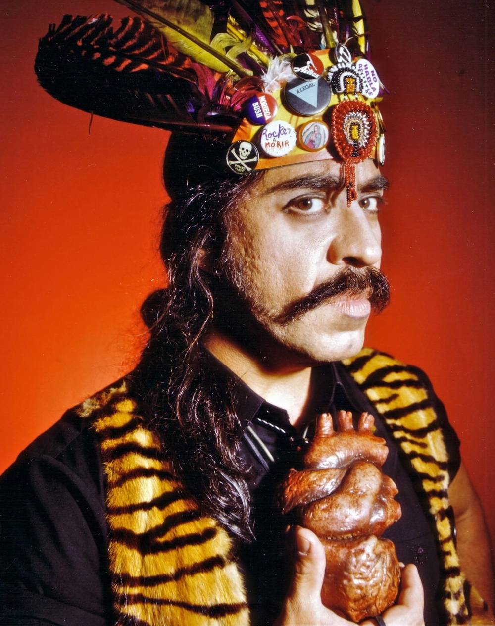 Portrait of Gómez-Peña in tiger-print vest, with feather headdress adorned with punk buttons, holding plastic replica of human heart.