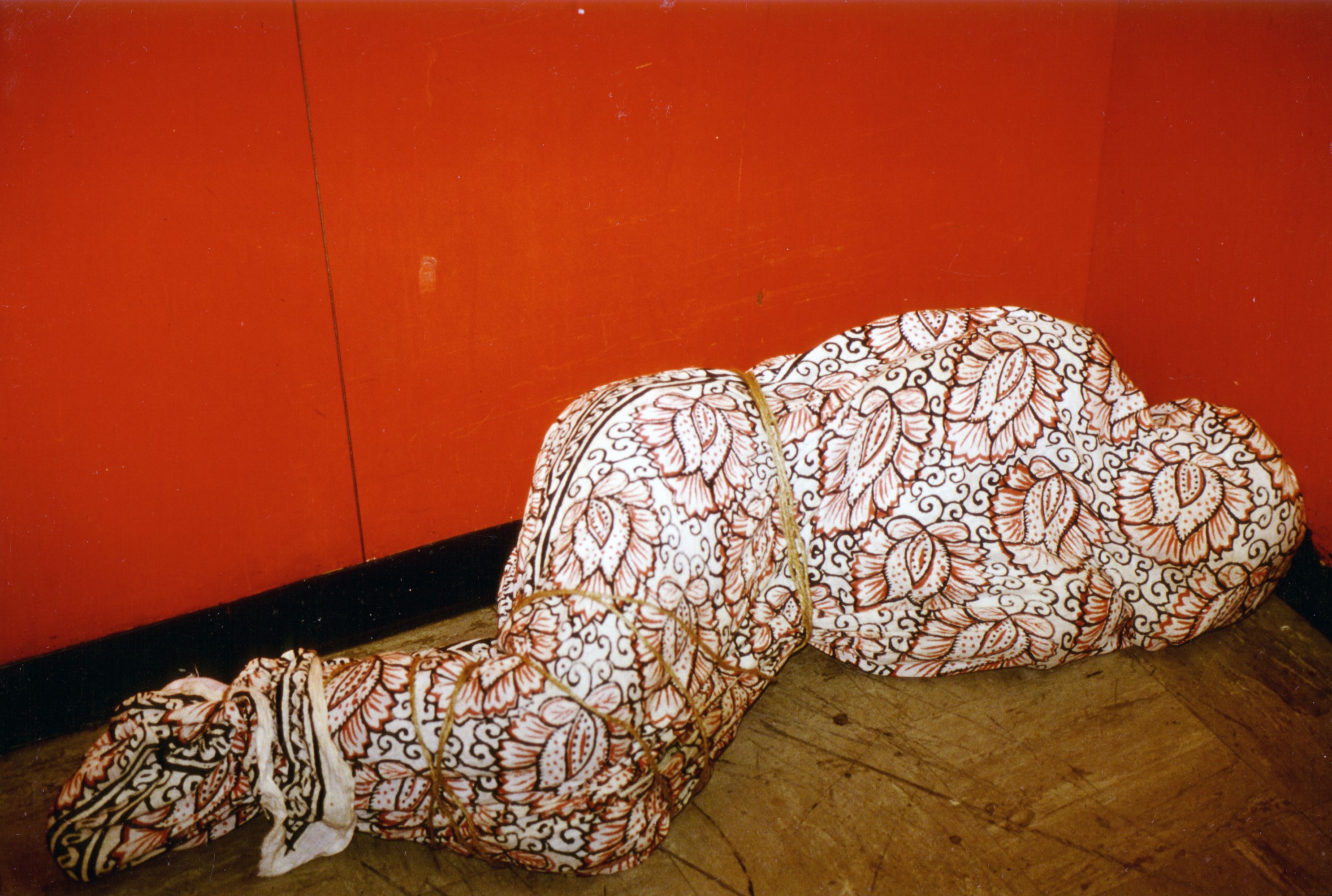 Guillermo Gómez-Peña positioned on the floor in a public elevator. Wrapped in floral-patterned batik fabric.