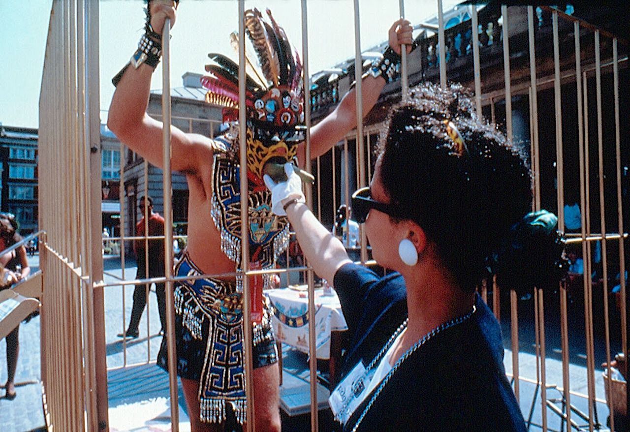 Woman in white gloves feeding Gómez-Peña inside the cage.