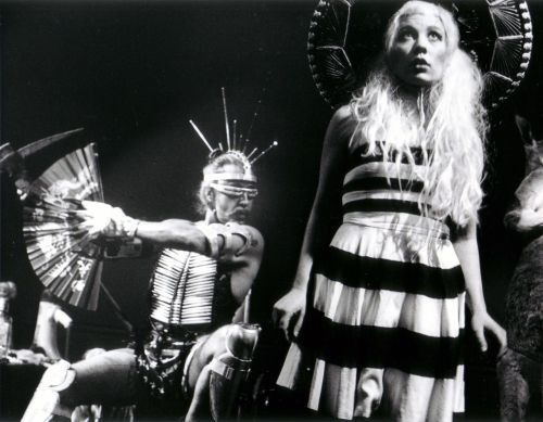 Guillermo Gómez-Peña seated with cyber-ethnic costumery and sunglasses next to a standing woman looking upwards with pale hair, sombrero, and a black and white dress.
