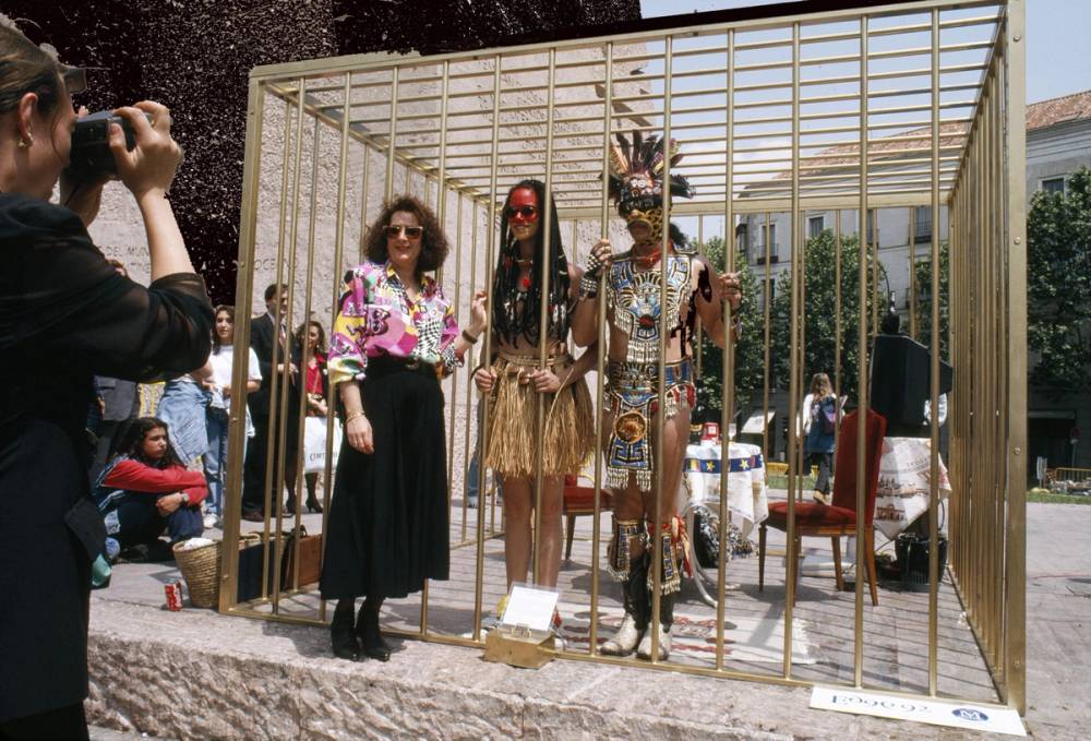 Lady posing next to to a golden cage with Gómez-Peña and Coco Fusco being exhibited within. 