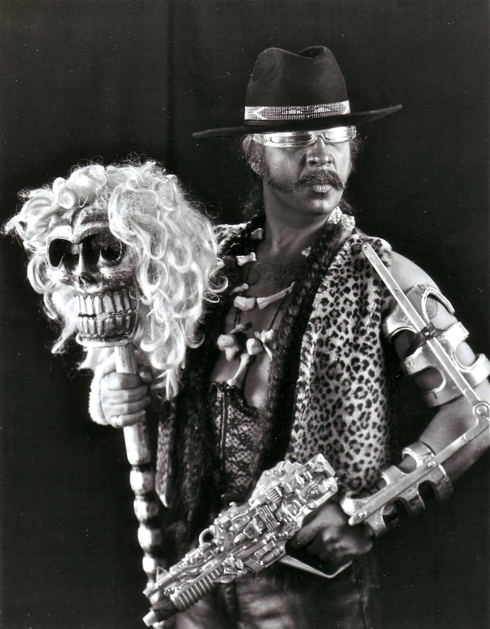 Guillermo Gómez-in sunglasses and a hat holding a toy gin and staff with a skull mask atop it.