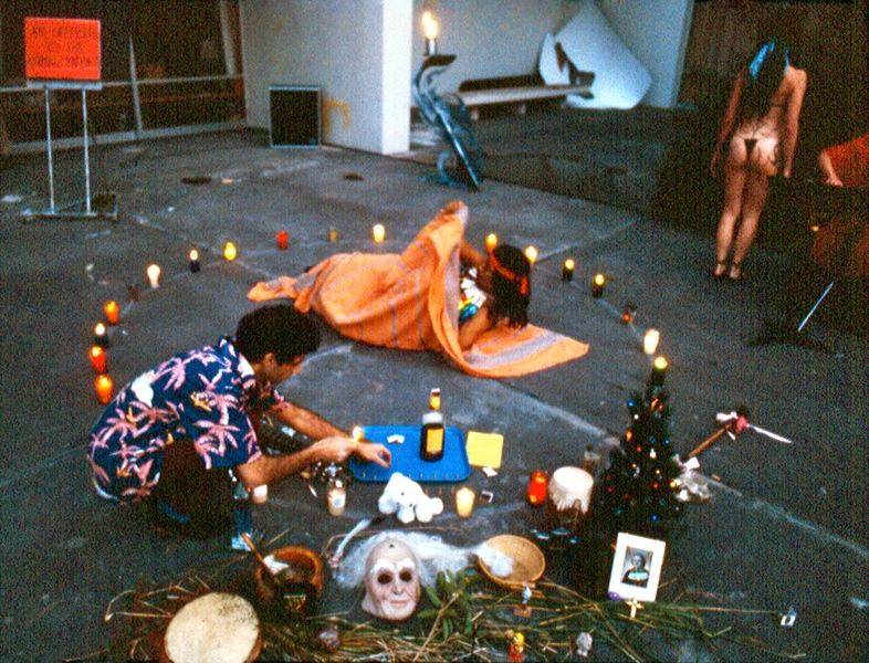 Guillermo Gómez-Peña crouching down in a ritual scene including candles, masks, photographs, and two other performers.