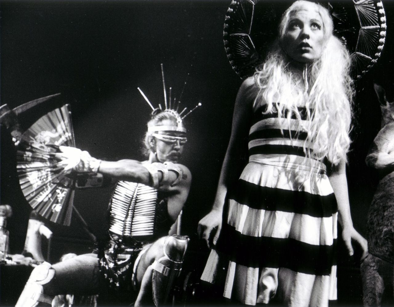 Guillermo Gómez-Peña seated with cyber-ethnic costumery and sunglasses next to a standing woman looking upwards with pale hair, sombrero, and a black and white dress.