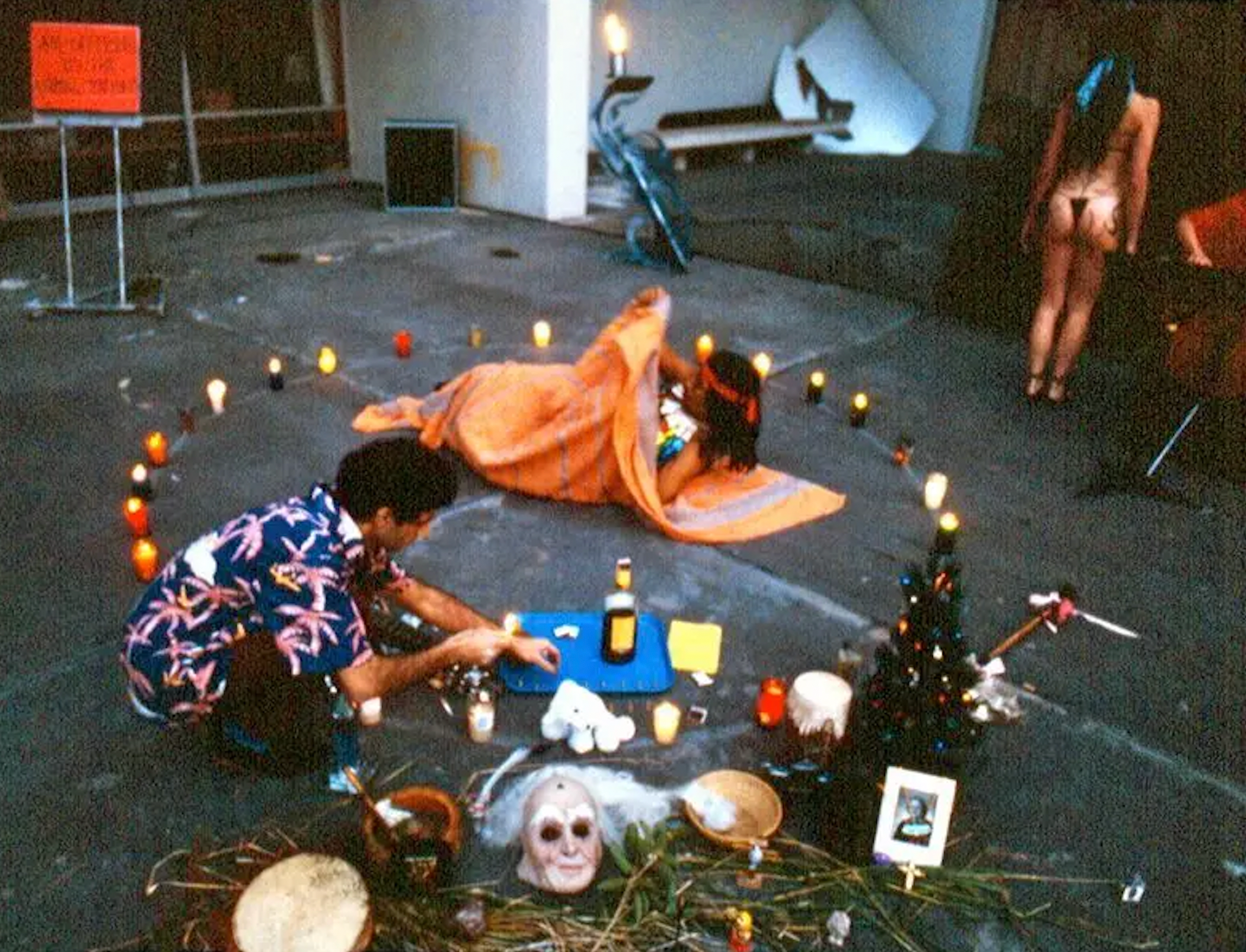Guillermo Gómez-Peña crouching down in a ritual scene including candles, masks, photographs, and two other performers.