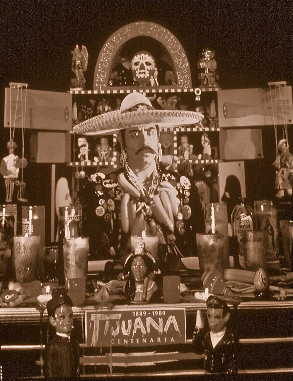 Guillermo Gómez-Peña sitting at an altar decorated with a kitsch collection of cultural fetish items, and wearing a border patrolman’s jacket decorated with buttons, bananas, beads, and shells.