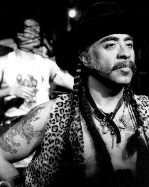 Guillermo Gómez-Peña in a leopard print vest, leather collar, and black hat. A figure in the background with bound face.