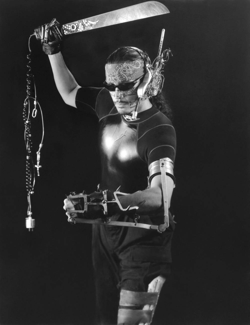 Roberto Sifuentes in latino cyborg garb. Machete in right hand, sunglasses, metallic prosthesis surrounding his left arm.