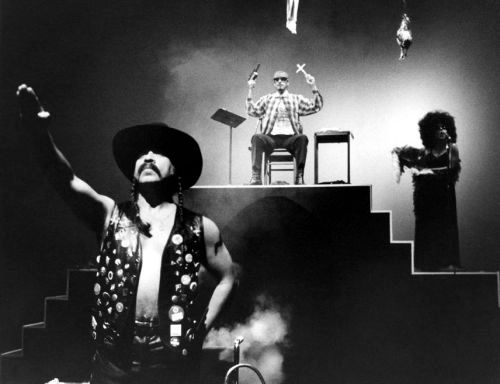Guillermo Gómez-Peña in a black leather vest and hat with right armed salute. Latino gang member on stage in the background with a woman in a dark dress nearby,