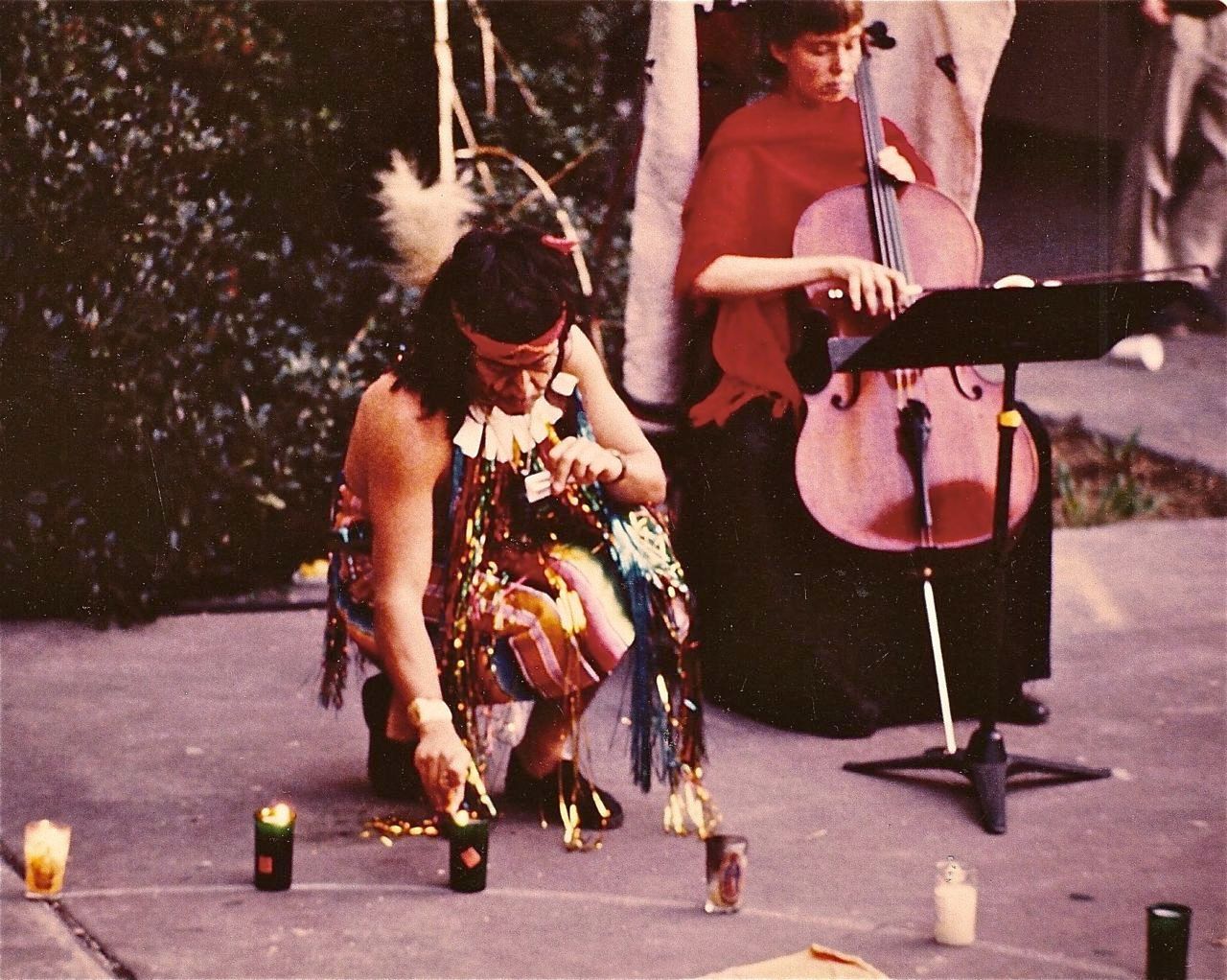 Guillermo Gómez-Peña lighting a ritual candle in shamanic attire. Female cellist playing in the background.