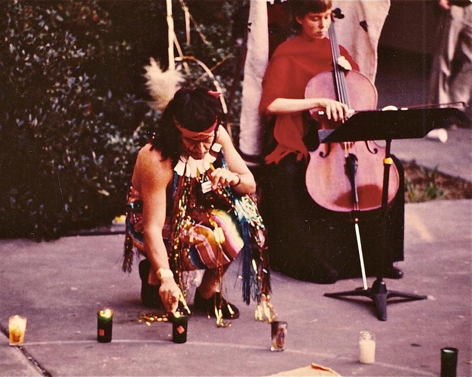 Guillermo Gómez-Peña lighting a ritual candle in shamanic attire. Female cellist playing in the background.