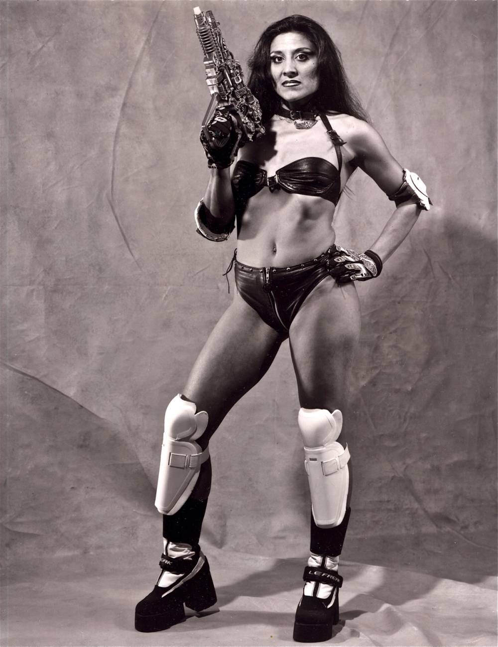 Latina woman in a bikini with a toy gun, knee and shoulder pads, high platform shoes.