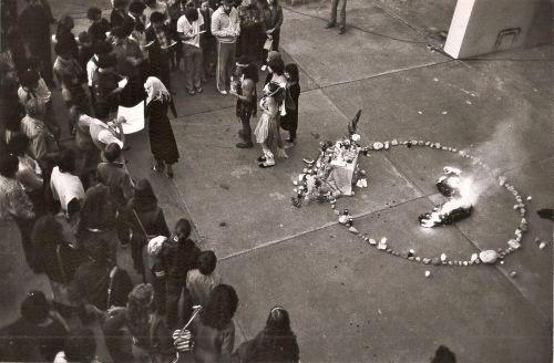 A crowd of people surround a circle of candles and ritual objects on a concrete floor.