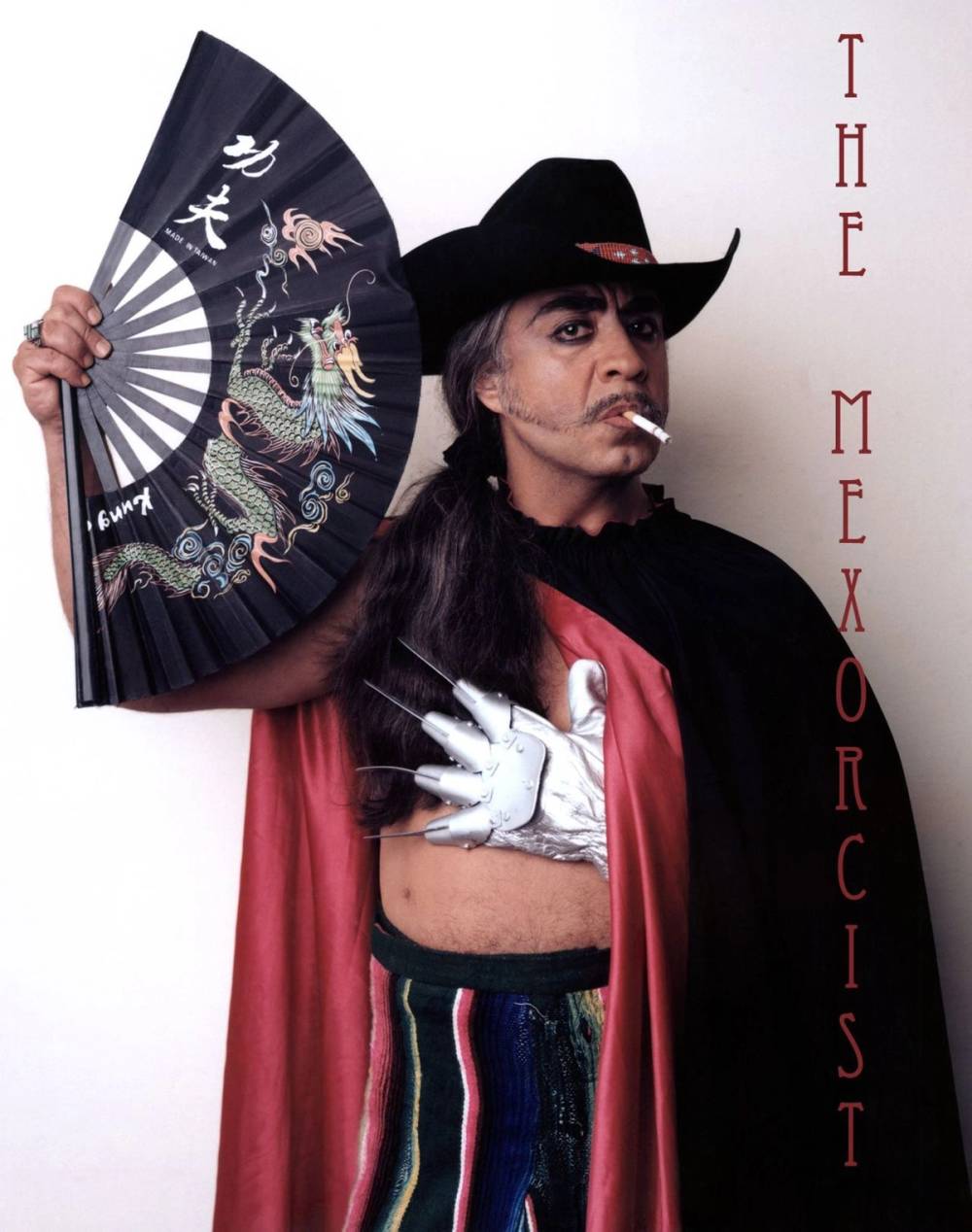 Gómez-Peña in black cape, black cowboy hat, holding an Asian fan in one hand and with a white Freddy Krueger style knife-glove in the other. 