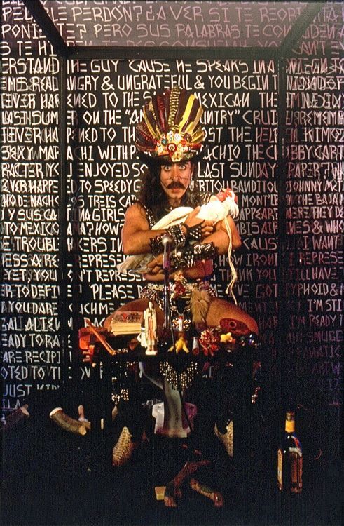 Guillermo Gómez-Peña seated inside a glass cabinet holding a white chicken, and surrounded by ritual artifacts.