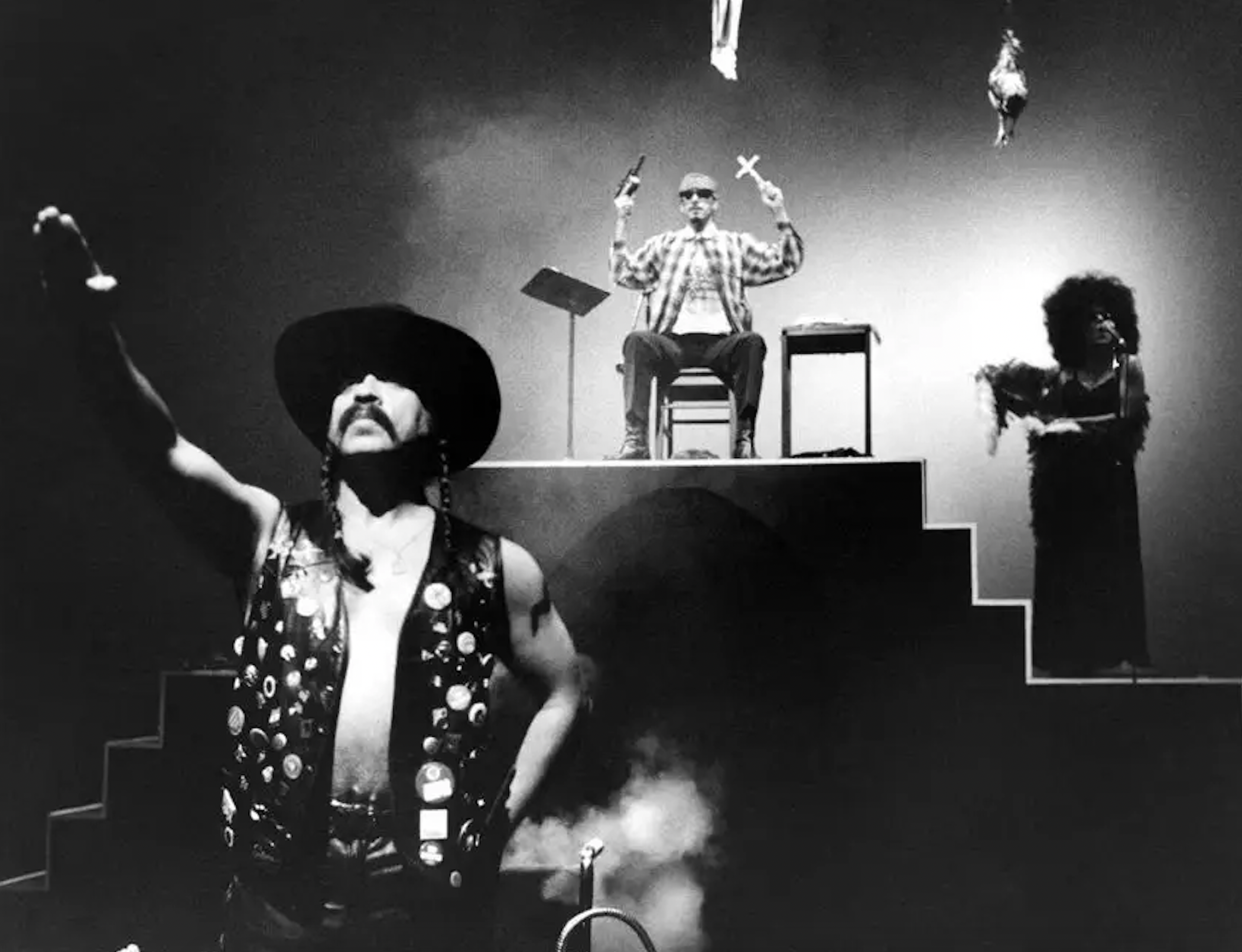 Guillermo Gómez-Peña in a black leather vest and hat with right armed salute. Latino gang member on stage in the background with a woman in a dark dress nearby,