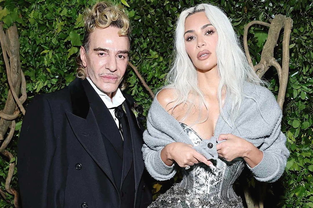 John Galliano in a black formal coat stands beside Kim Kardashian