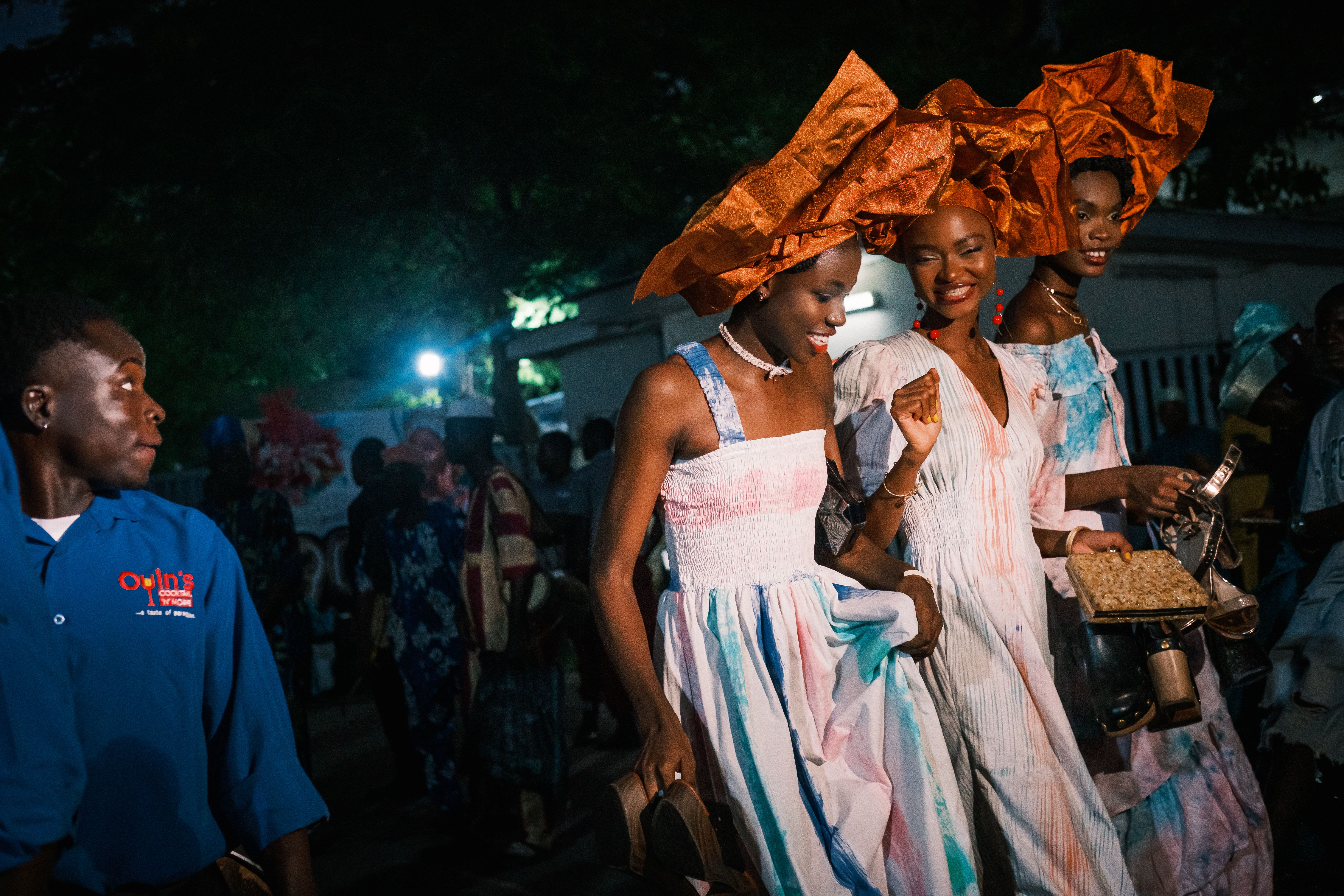Image placeholder. Three models in copper geles and pale tie-dyed gowns dancing inside the tent, clutches in hand, seated guests in blue behind them.