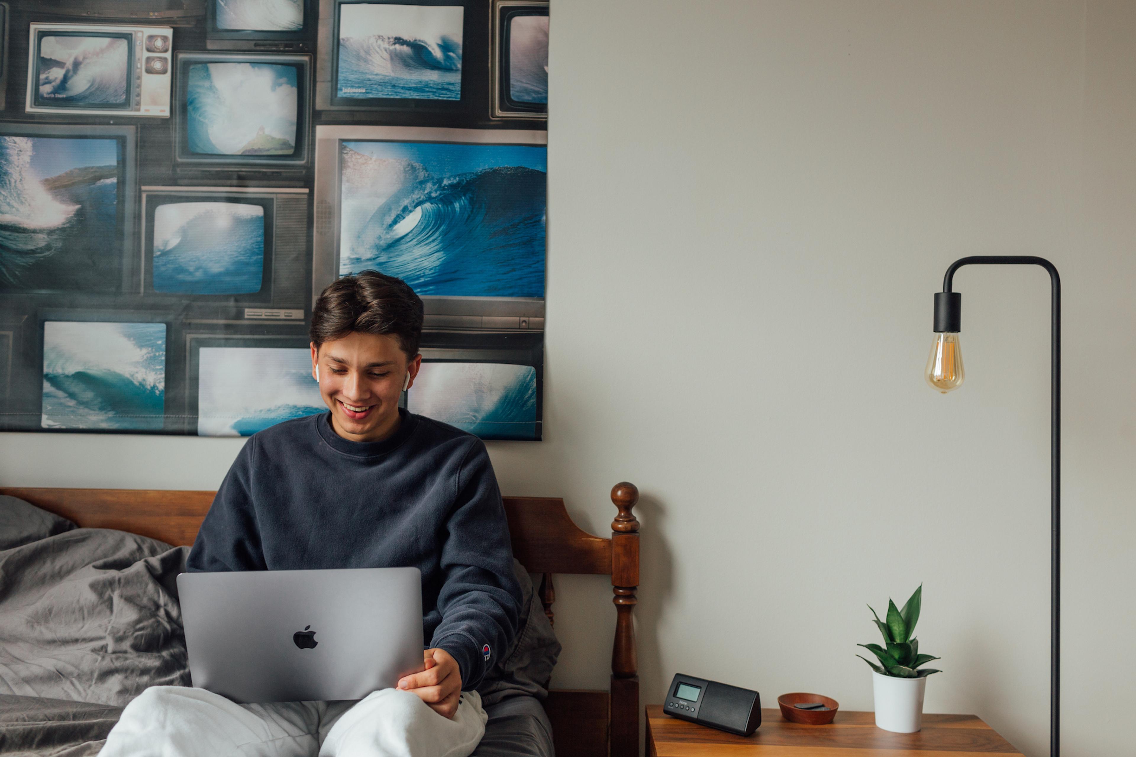 Young man smiling looking at a computer while sitting on a bed.