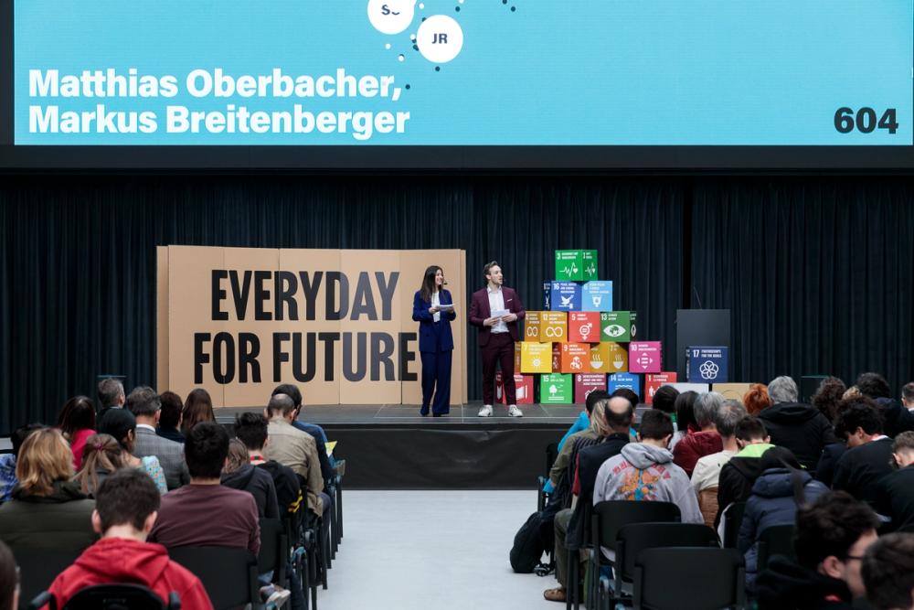 presentation stage with two speakers, big screen in background, audience visible in foreground