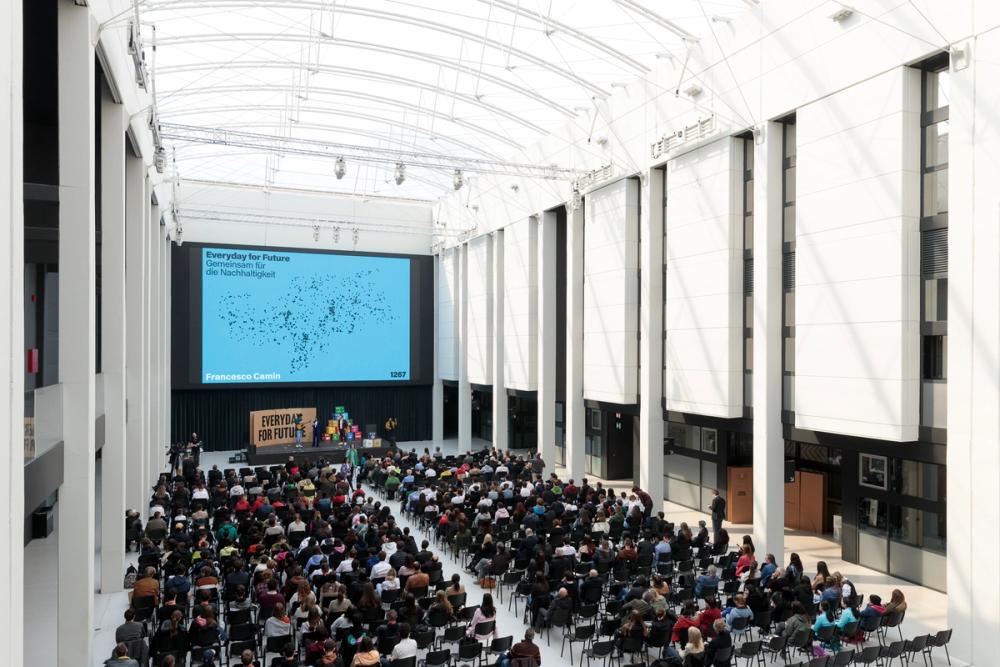 Large hall filled with people sitting and looking at a stage and and large presentation screen