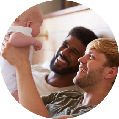 Two men are reclining next to each other. One is holding a baby above them; they are smiling at one another.