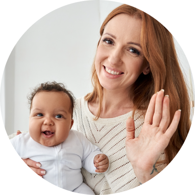 Smiling woman holding a baby, waving at the camera. 