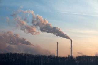 Three industrial smokestacks emitting large plumes of smoke into the sky above an industrial area. Bare trees are visible in the foreground, and the scene is tinted in blue and purple tones, illustrating industrial emissions and air pollution.