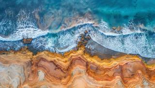Ariel view of rocky coastline with colourful sand beside ocean with waves 