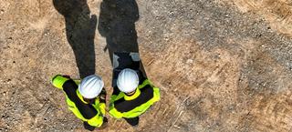 Aerial image of two workers in high-vis and hard hats looking at a laptop and the ground, which is bare soil