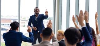 Presenter at the front of the room while seated people raise their hands