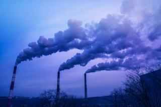 Three industrial smokestacks emitting large plumes of smoke into the sky above an industrial area. Bare trees are visible in the foreground, and the scene is tinted in blue and purple tones, illustrating industrial emissions and air pollution.