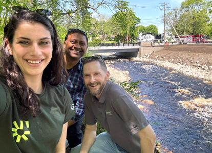 Taylor DelVecchio and Andie Greene of Water Resources Engineering and Kishor Patel of Structural Engineering are pictured in a selfie, posed in front of the High Street bridge over the Town River.