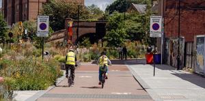 Two people in fluorescent jackets riding their bikes on a bike path in a city, next to some flower beds
