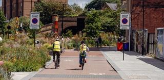Two people in fluorescent jackets riding their bikes on a bike path in a city, next to some flower beds