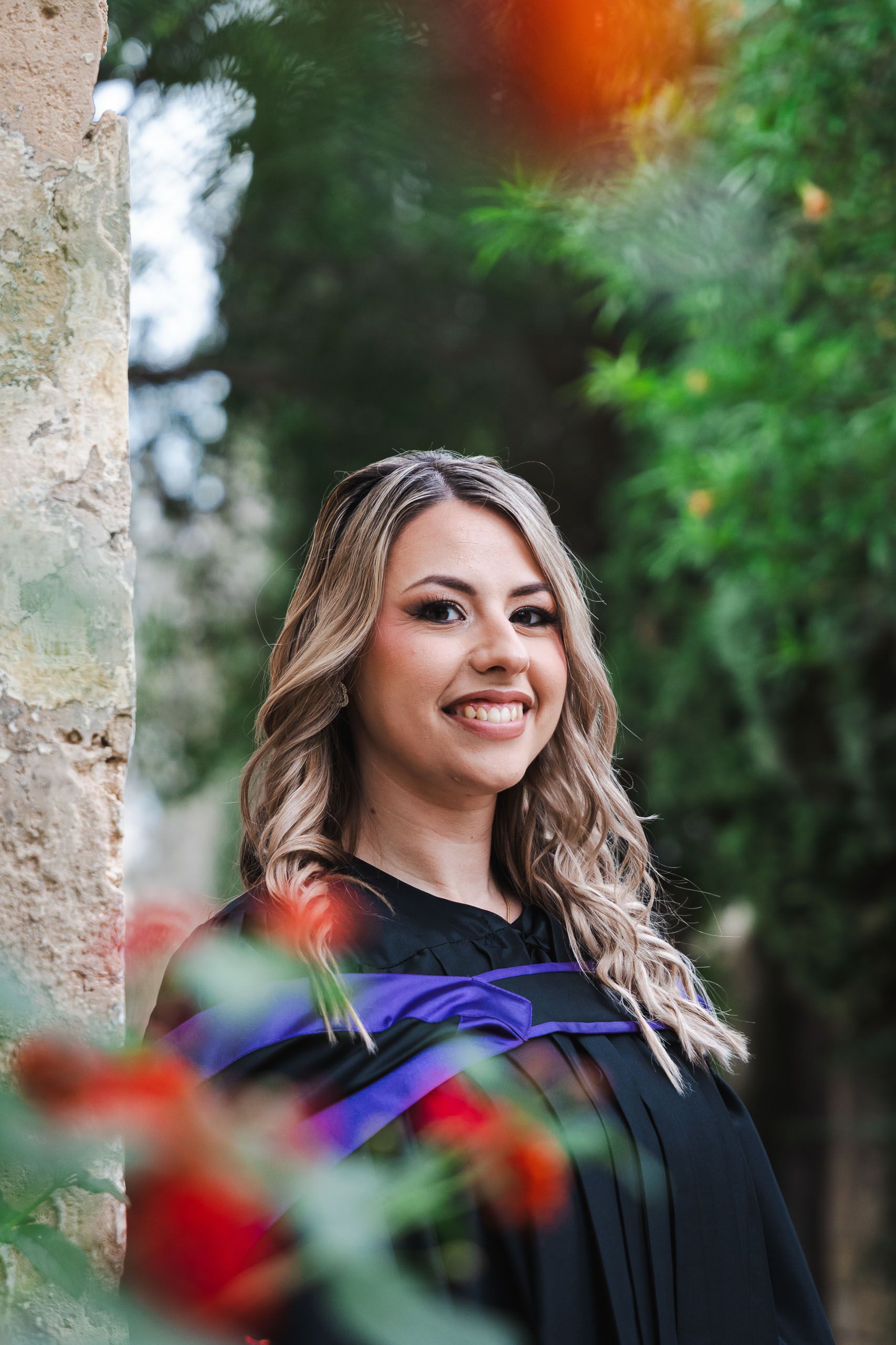 Smiling graduate in a purple-trimmed gown photographed through red flowers at San Anton Gardens in Malta.