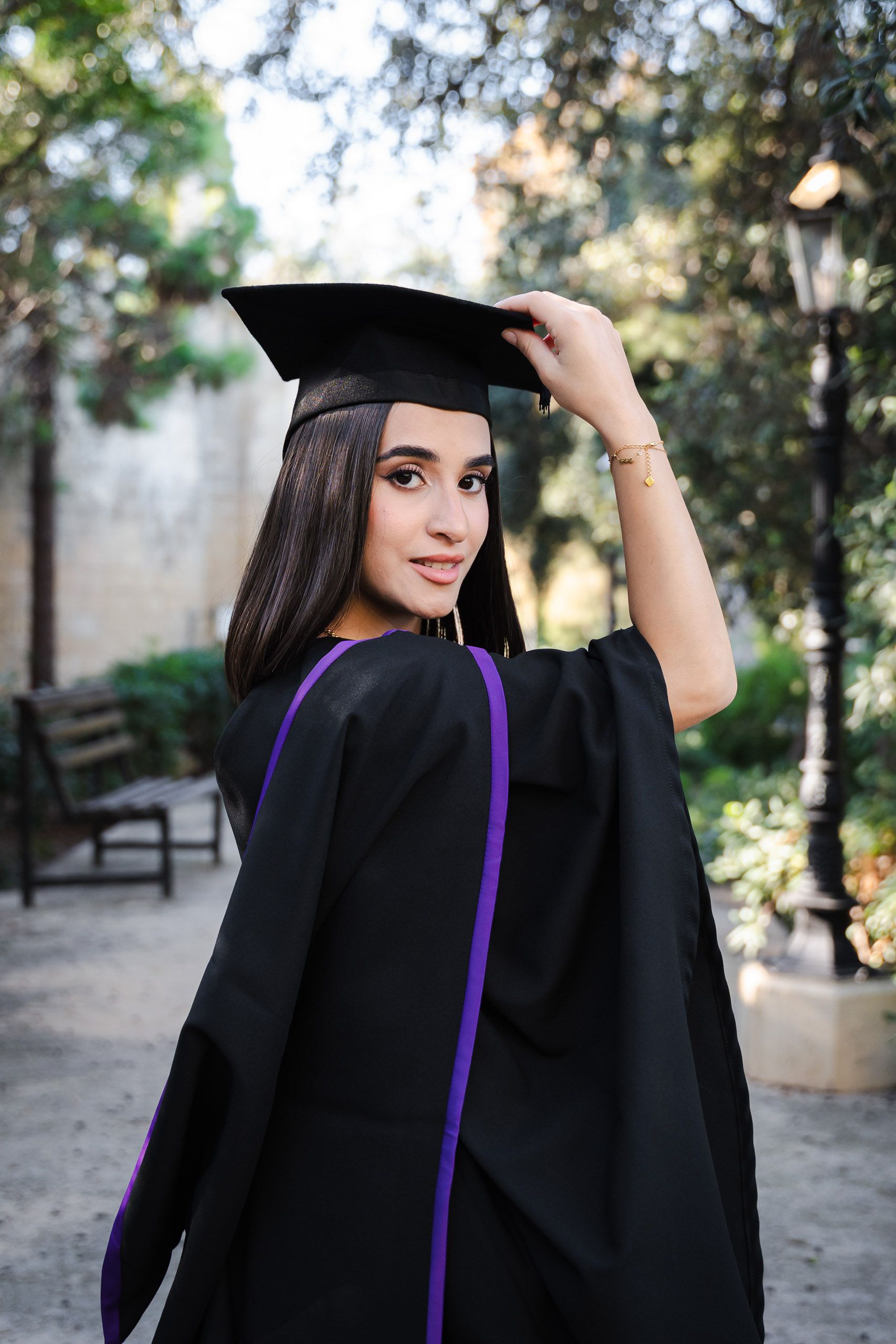 Graduate wearing a cap and purple-trimmed gown looking back at the camera at Romeo Romano Gardens in Malta.